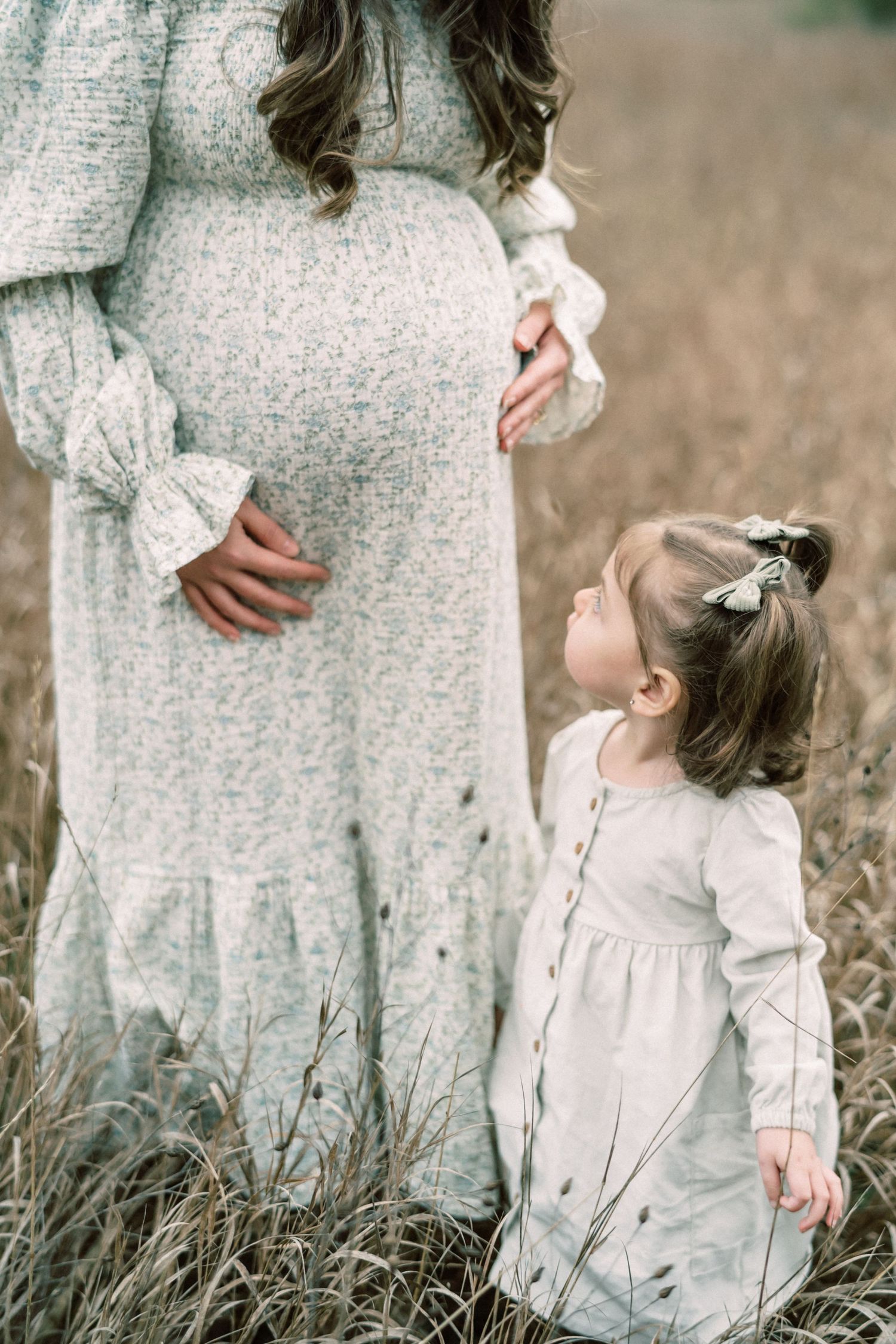 Expectant mother in grey dress stands with young child in matching dress among tall grass.