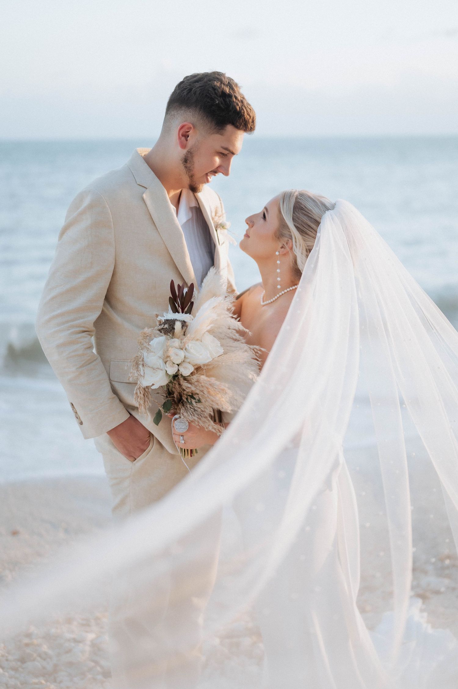 A bride and groom share an intimate moment on a beach at sunset, with her long veil flowing in the breeze.