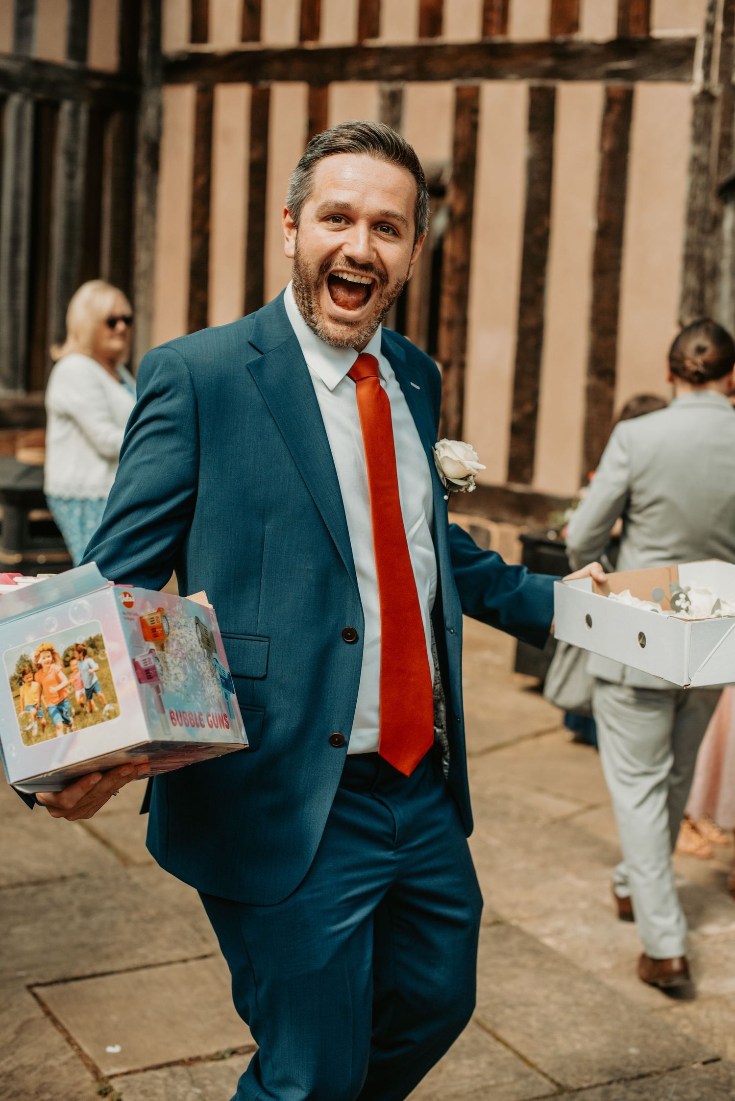 A person in a navy business suit and red tie laughs joyfully while holding decorated boxes on a city street.