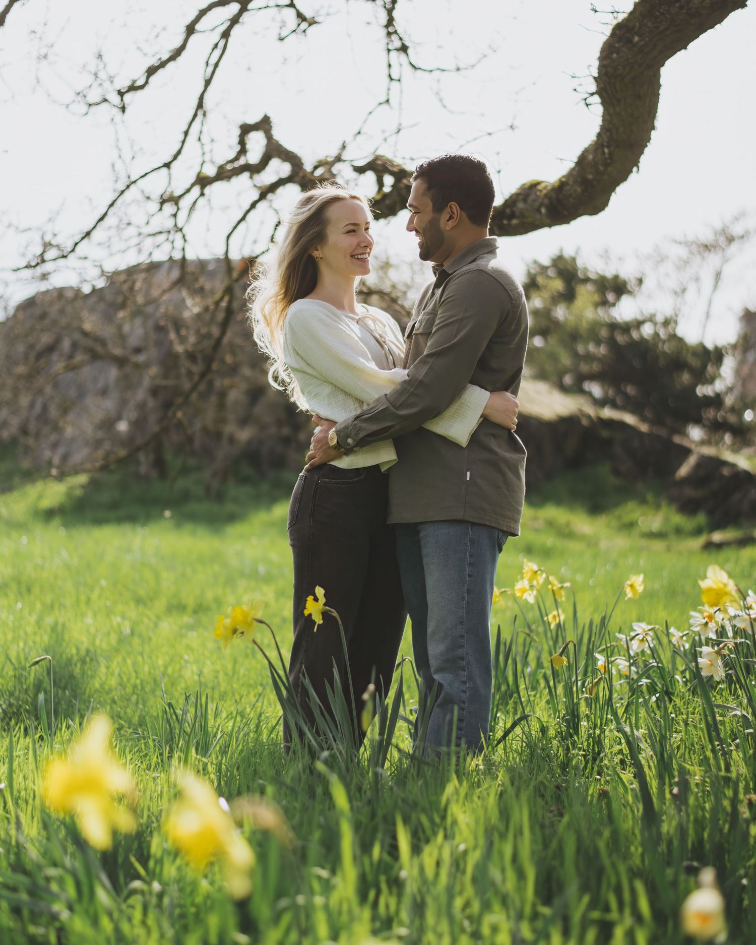 Two people embrace while standing in a green meadow under a gnarled tree branch on a sunny spring day.