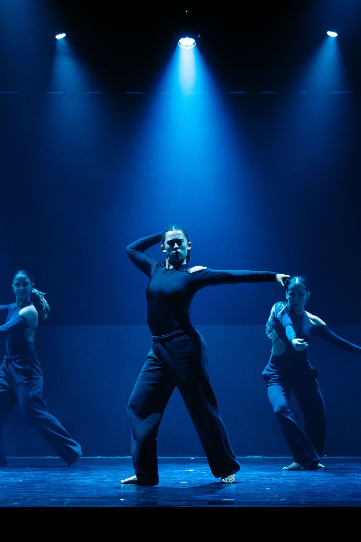 Modern dancers in black attire perform under dramatic blue spotlights on a dark stage.