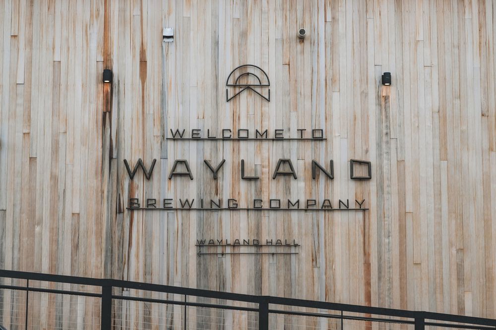 Welcome to Wayland Brewing Company sign mounted on light wooden wall with black metal railing in foreground.