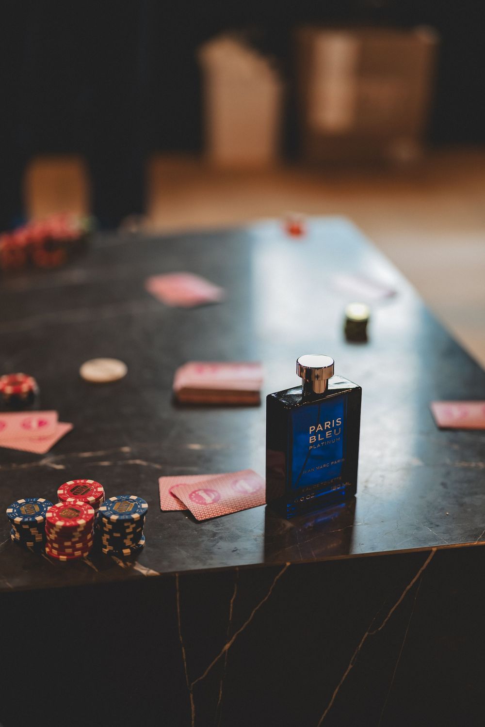 Playing cards and poker chips scattered on a dark table with a bottle of Paris Blue perfume.