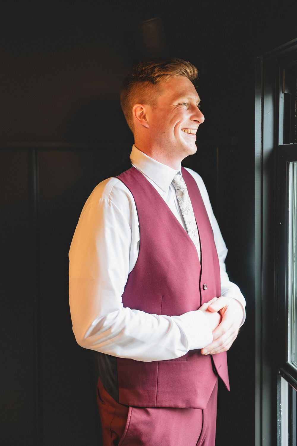 Groom in burgundy vest and white dress shirt smiling while standing near a window.