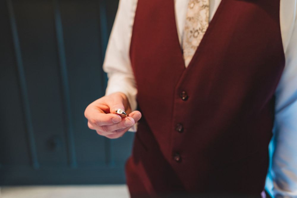 Close-up of hands holding wedding rings while wearing burgundy vest.