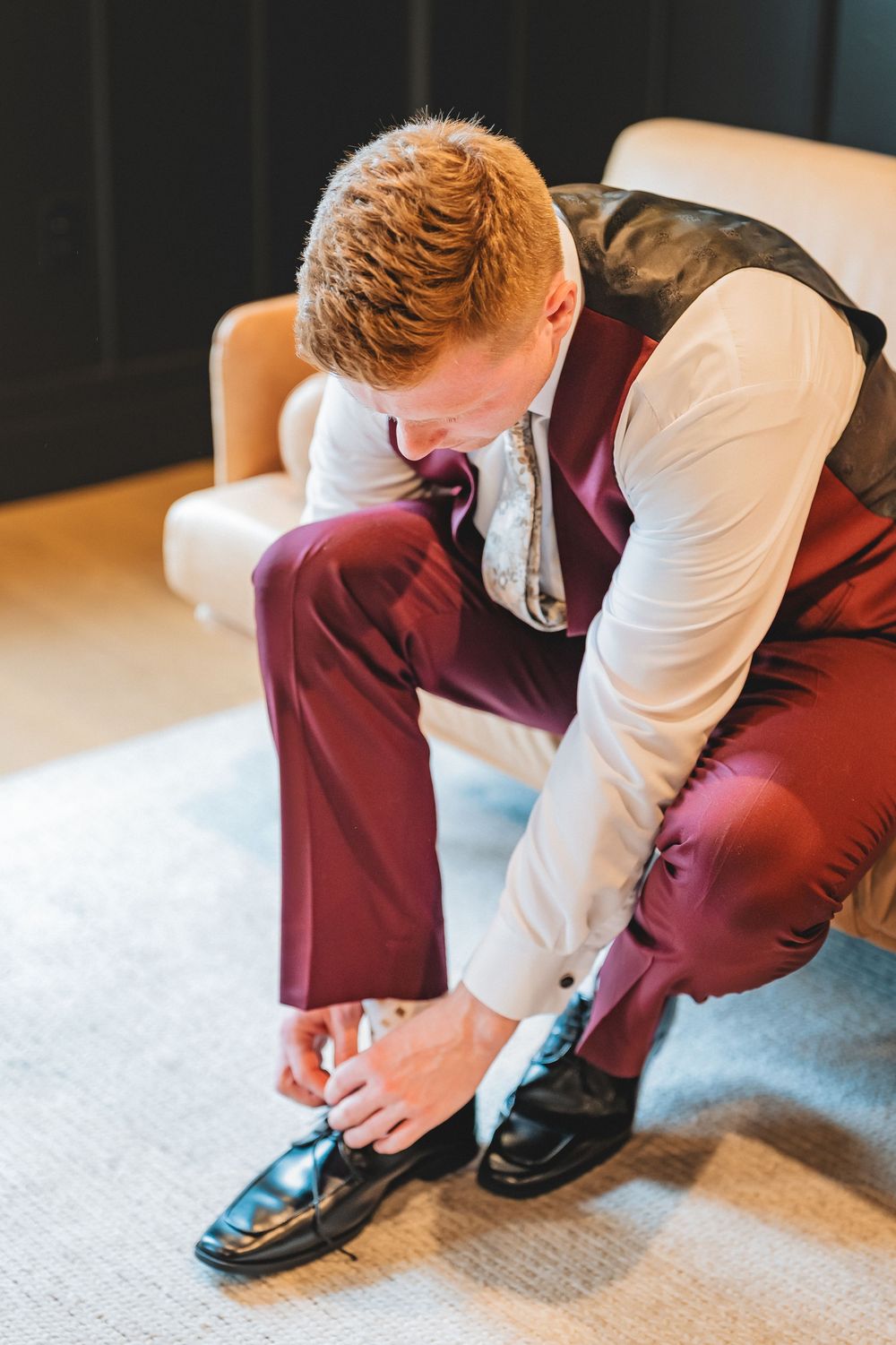Groom in burgundy suit and white shirt tying formal dress shoes while sitting on light gray carpet.