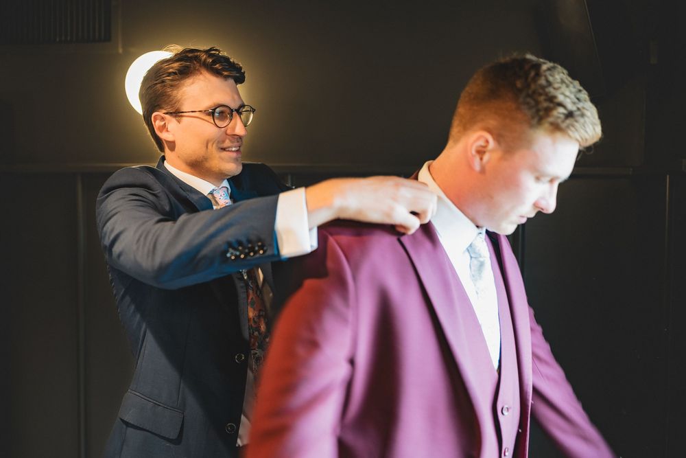 A man in a navy suit helps adjust the clothing of another person wearing a pink suit jacket.