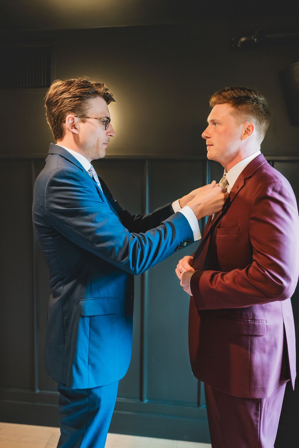 Two people in suits, one navy and one burgundy, share a formal moment as one adjusts the other's tie.