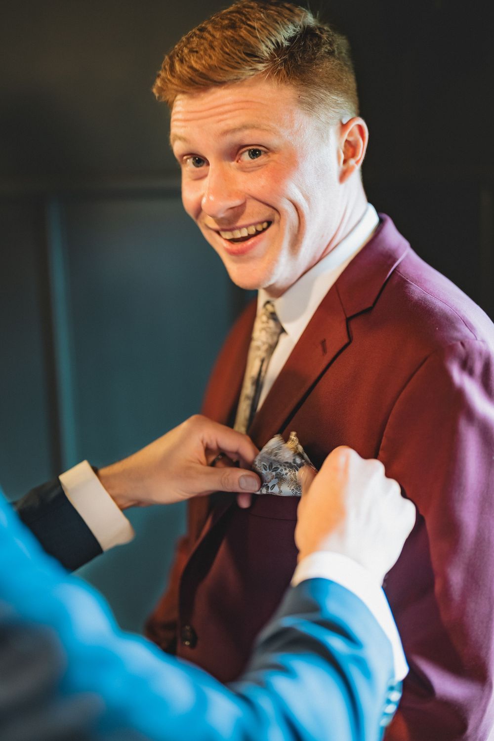 A groom in a burgundy suit smiles while someone adjusts his boutonniere before the wedding ceremony.