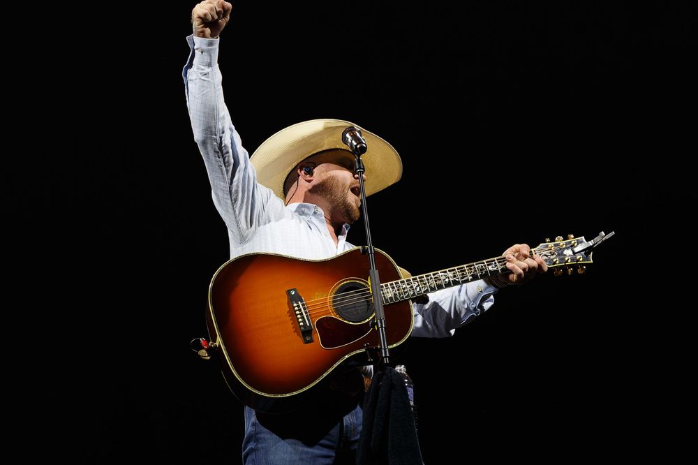 Country music performer Cody Johnson in cowboy hat playing acoustic guitar on stage with dramatic lighting at Bridgestone Arena.