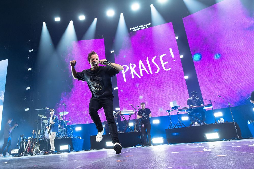 Performer for Elevation Worship jumping on stage with 'PRAISE!' text displayed on purple backdrop during concert at Bridgestone Arena.
