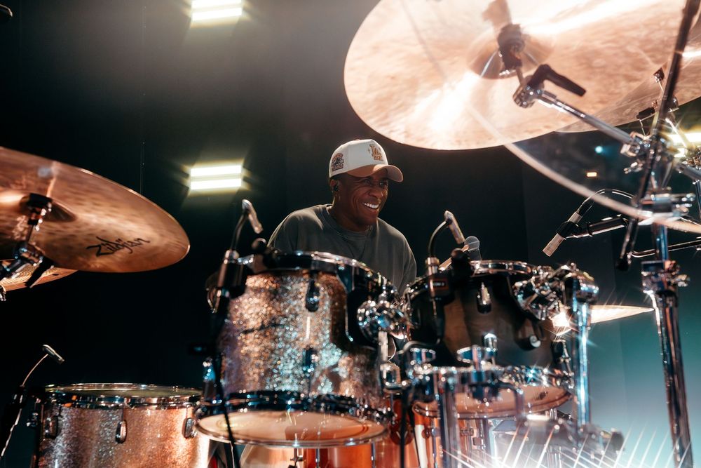 Drummer performs behind extensive drum kit setup with dramatic overhead cymbal lighting.