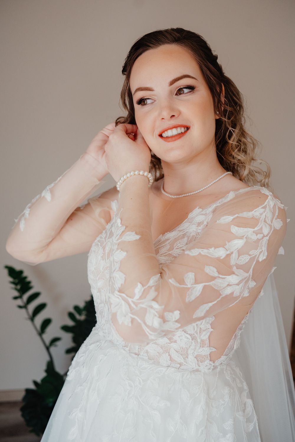 A bride wearing a romantic white lace wedding gown with sheer sleeves and pearl accessories smiles while looking to the side.
