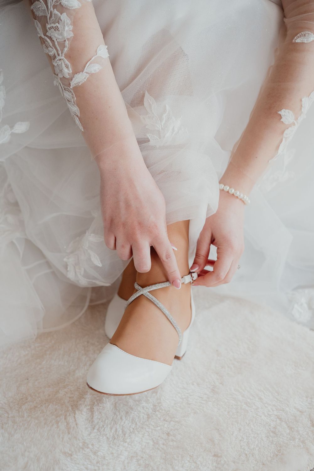 Hand fastening white wedding shoe strap against tulle dress fabric and light carpet background.
