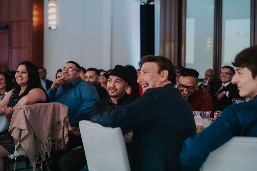 A crowd of attendees sits in white chairs during an indoor event, listening attentively in a warm-lit venue.