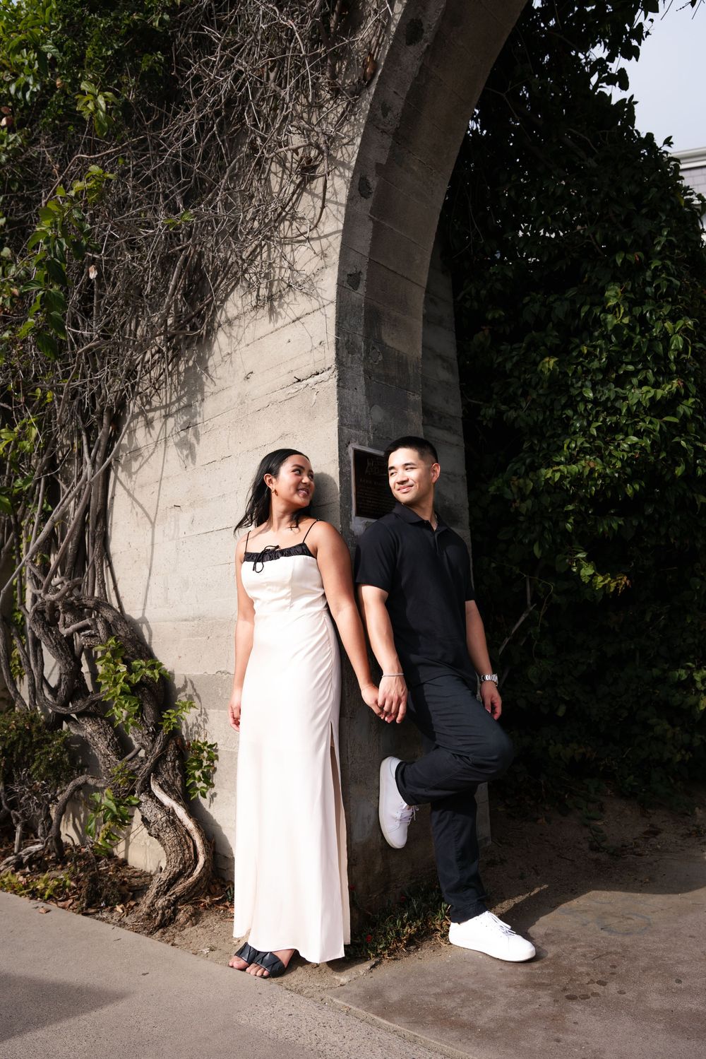 Couple in casual and formal attire standing together under a stone archway with climbing vines during sunset.