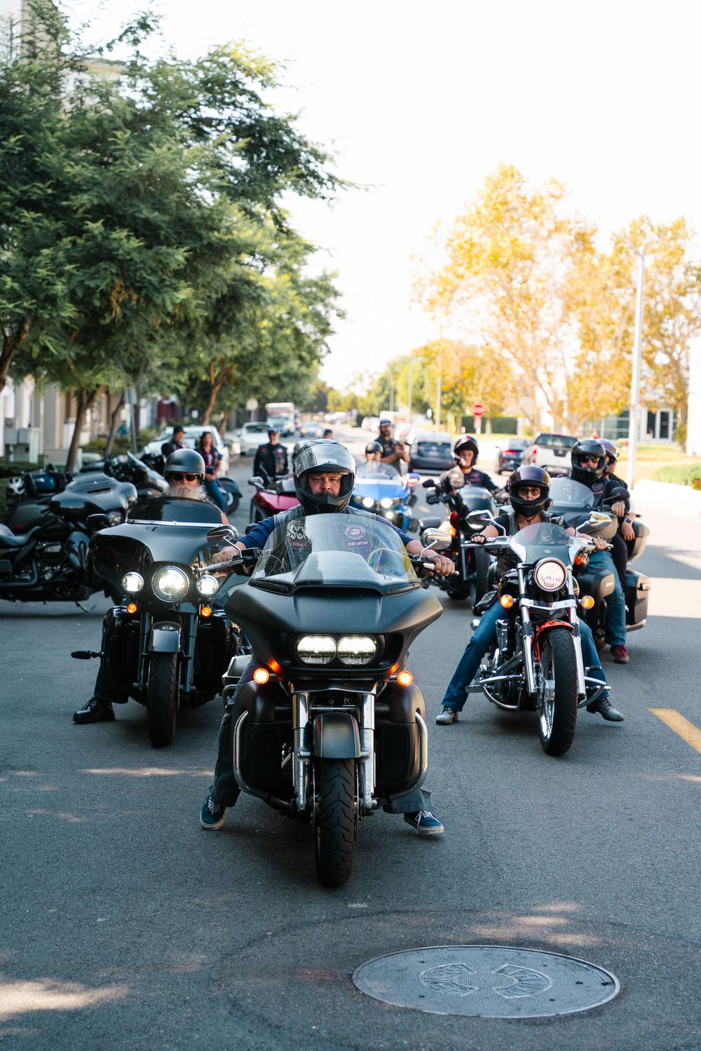 A group of motorcycles and riders gathering on a tree-lined street during daytime with their headlights on.