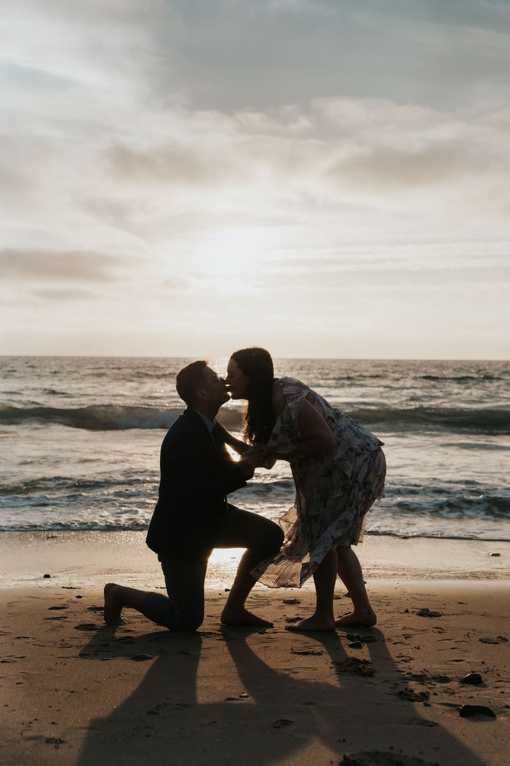 A romantic silhouette proposal scene on a beach at sunset with waves and cloudy skies in the background.