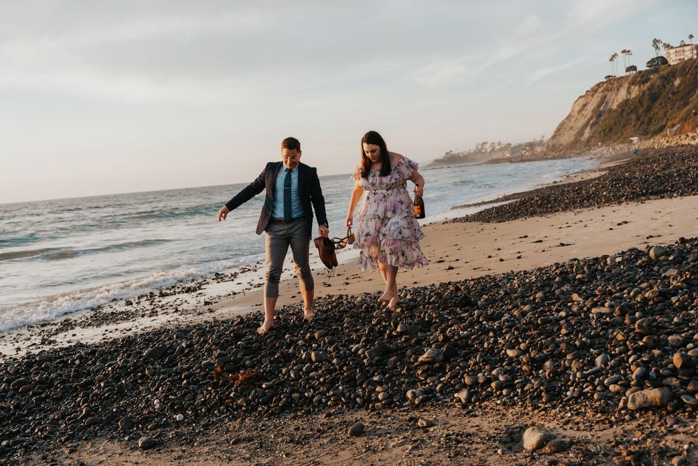 A couple walks hand in hand along a rocky beach at sunset with coastal cliffs in the background.