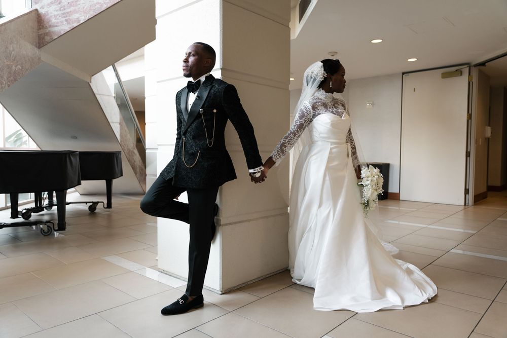 Newlywed couple holding hands while standing on opposite sides of a wall near a piano in an elegant venue.