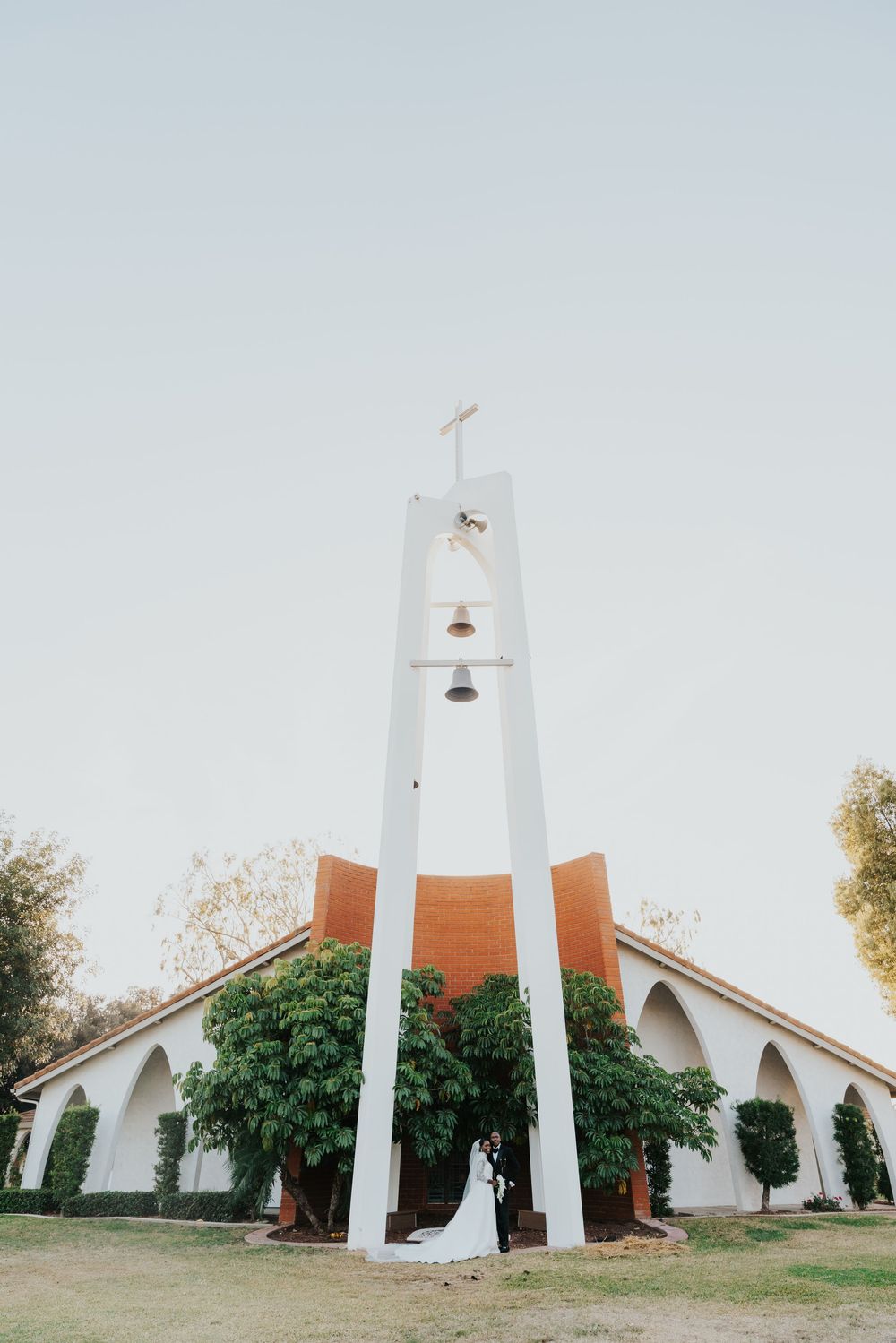 Modern white church steeple with cross and bells against pale blue sky at wedding venue.