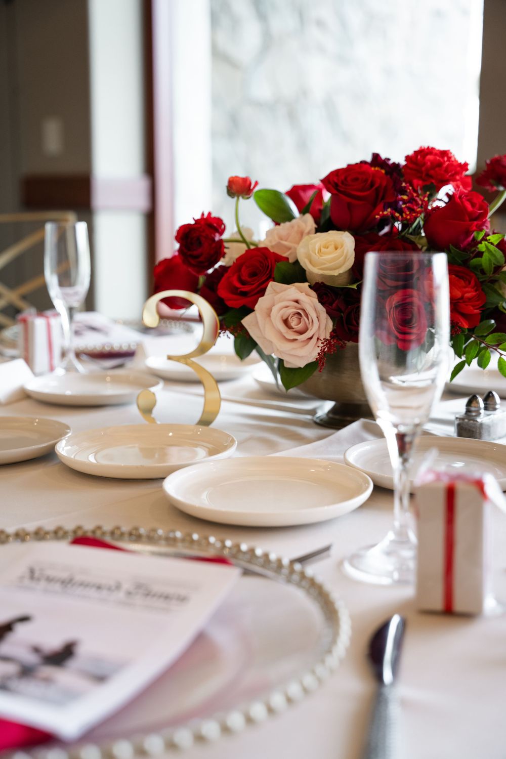 Elegant table setting with red and pink roses centerpiece, crystal glasses, and gold table number at a formal event.
