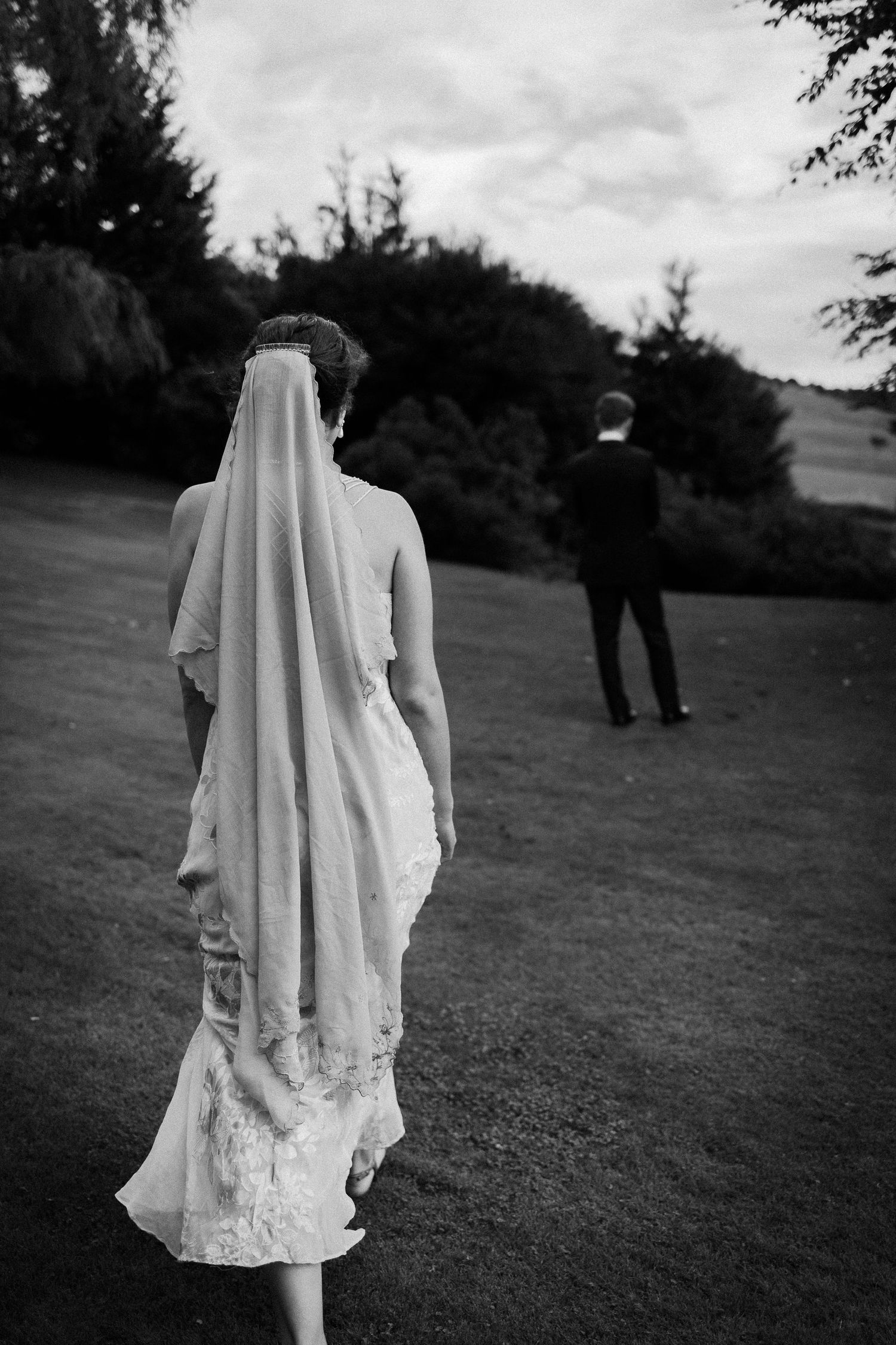 Black and white wedding photo showing a bride in a flowing veil and gown approaching a distant groom on grassy grounds.