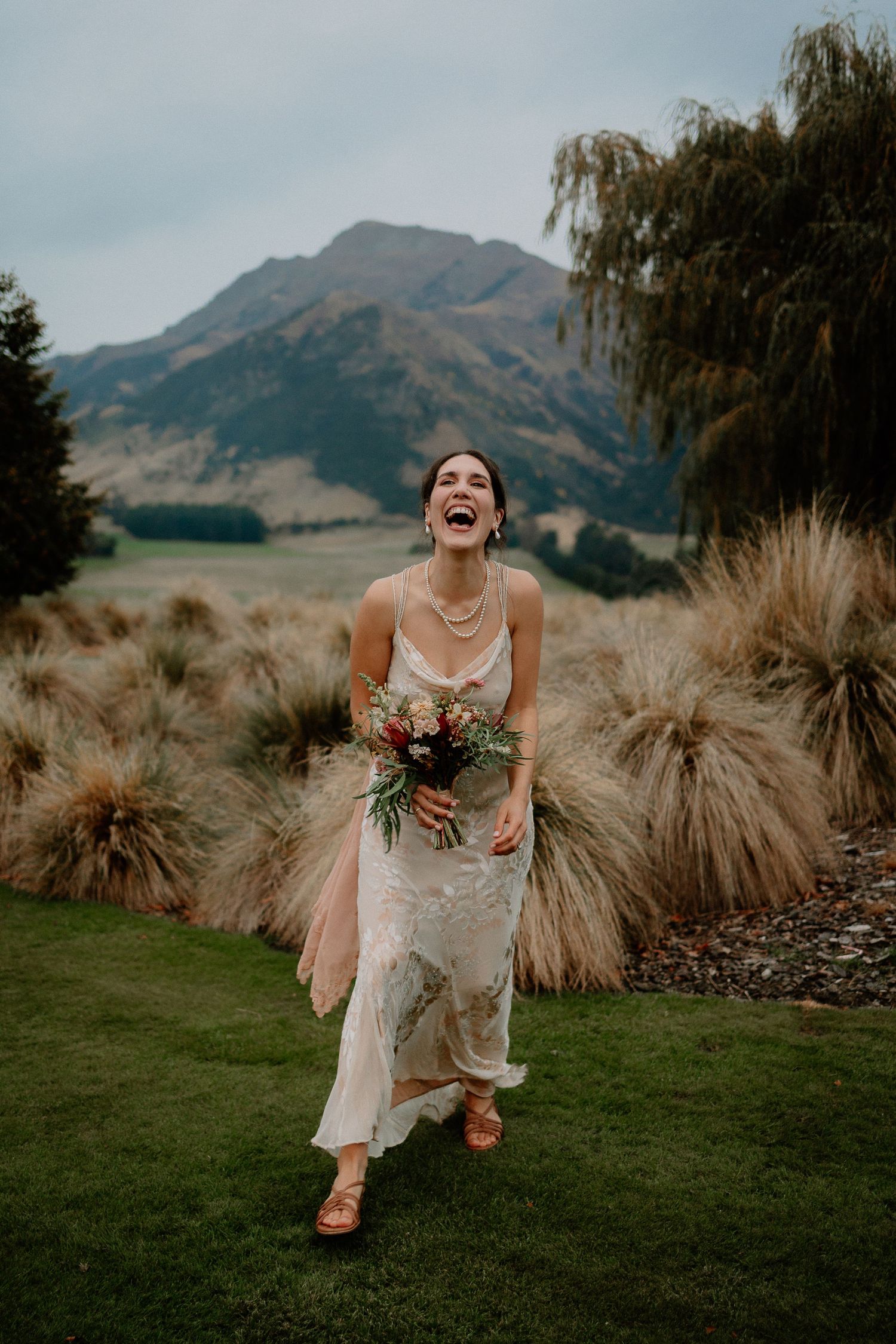 Bride in lace wedding dress laughs joyfully against mountain backdrop with ornamental grasses and willow trees.