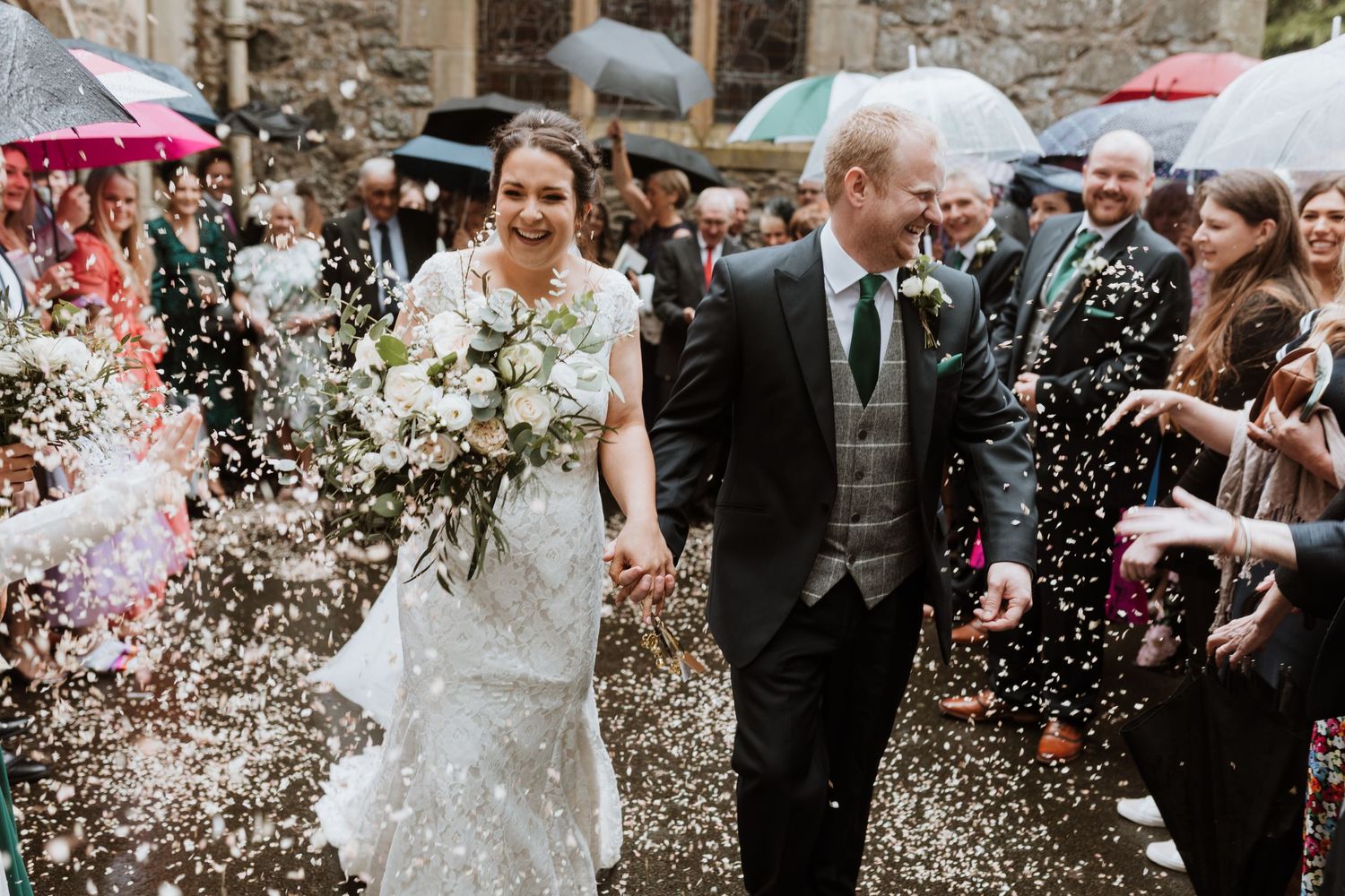 A joyful wedding couple walks through confetti as guests throw petals at them outside a church in the rain.