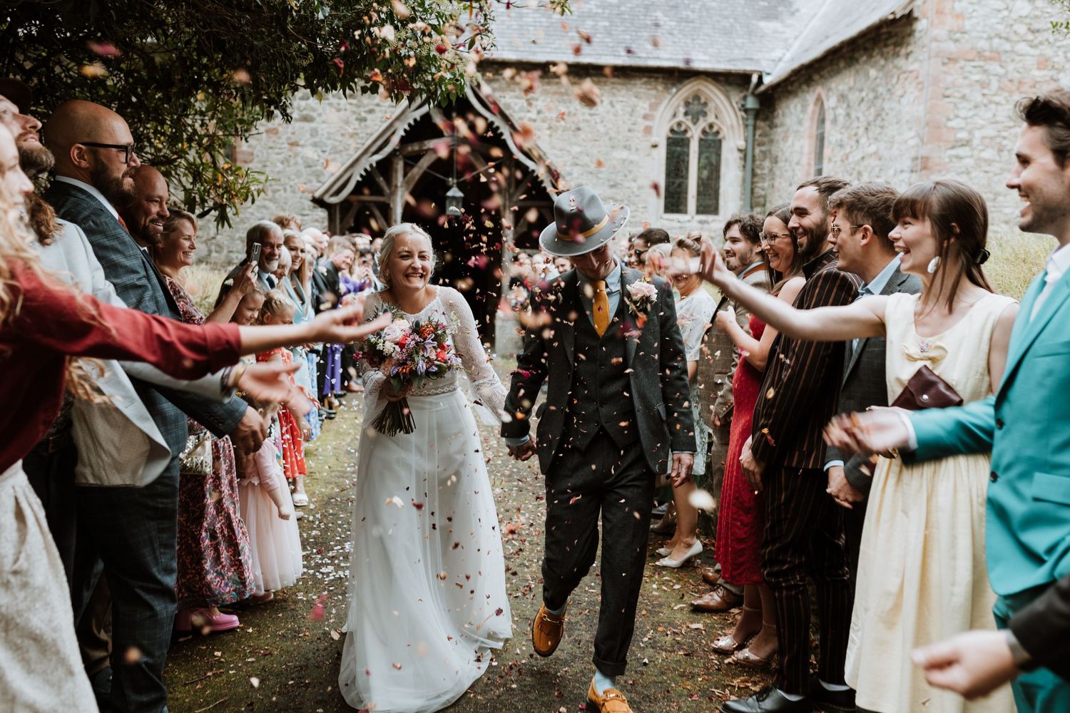 Wedding guests toss confetti over a wedding couple exiting a church ceremony while walking through a crowd.