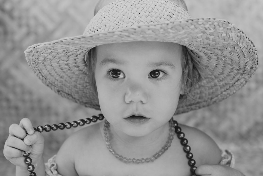 A black and white portrait of a small child wearing an oversized straw sun hat and beaded necklace.