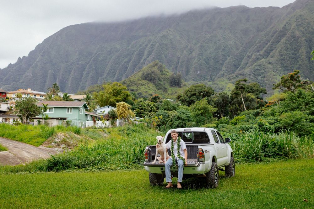 Two people loading a white pickup truck in a lush green valley with dramatic mountain cliffs in Hawaii.
