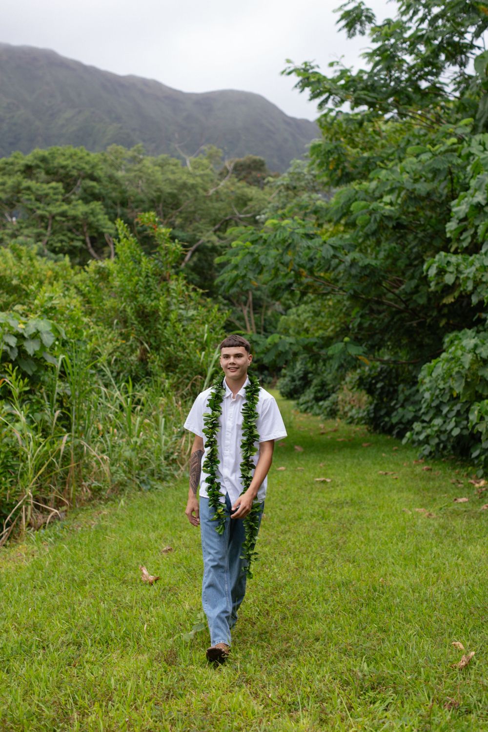 A person wearing a white floral lei walks along a grassy path surrounded by lush tropical vegetation and mountains.
