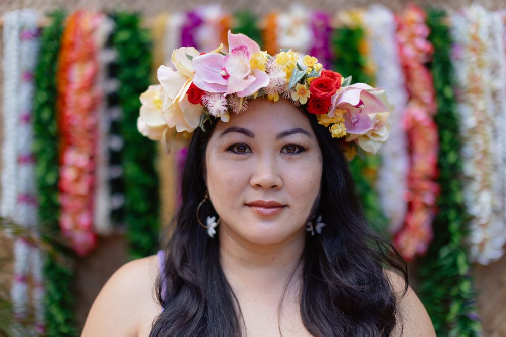 A floral crown with pink, yellow and red blooms adorns dark hair against a colorful backdrop of hanging decorations.