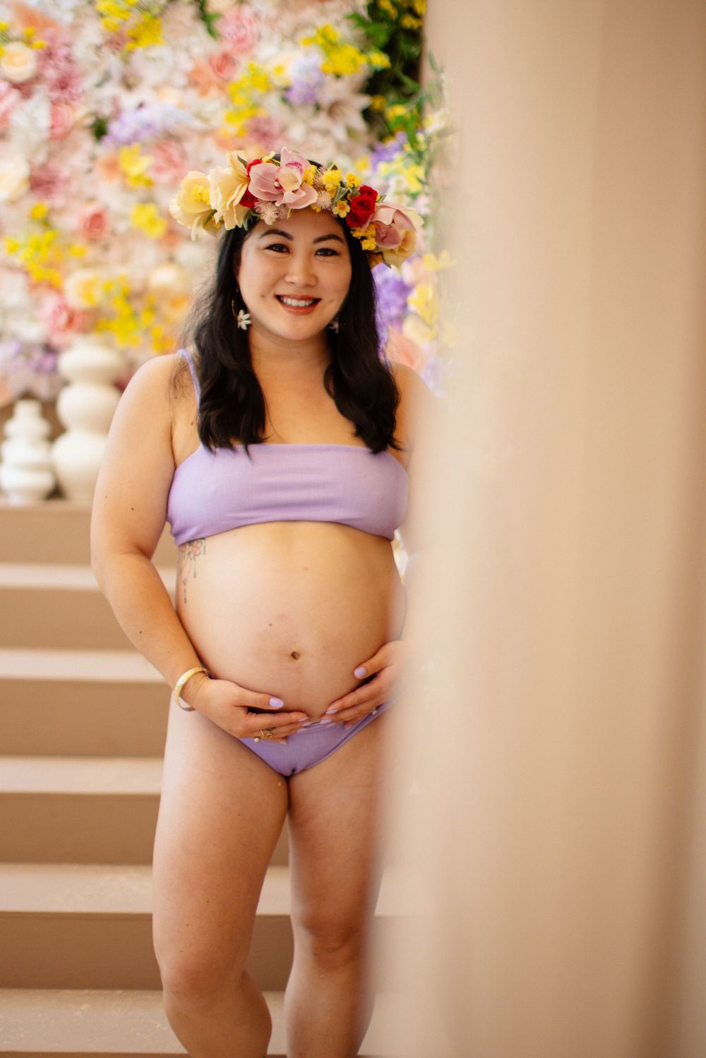 A pregnant person wearing a purple swimsuit and floral crown poses on stairs with floral decorations in the background.