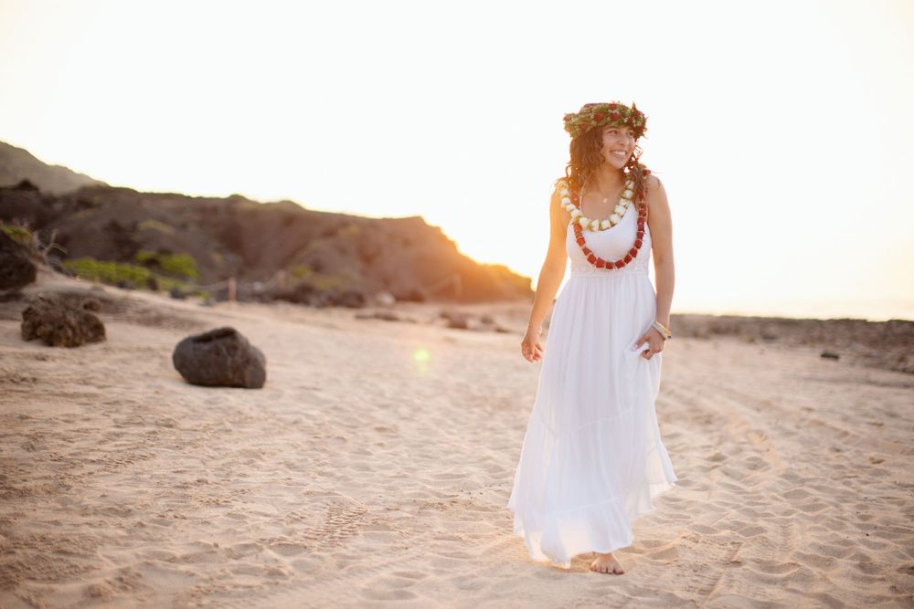 A person in a flowing white dress with a red lei necklace walks on a sandy beach at sunset.