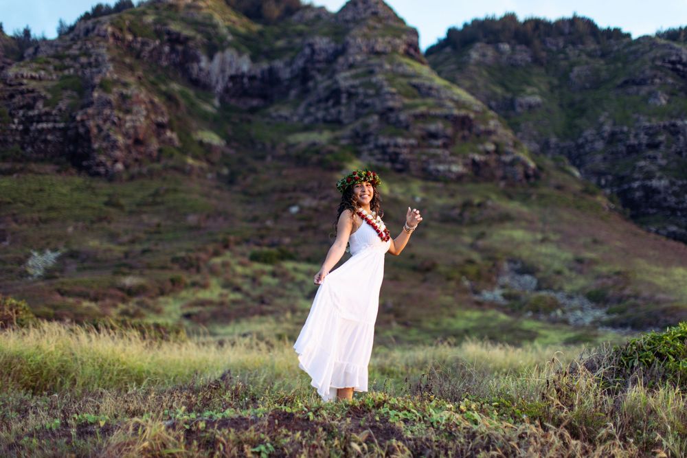 A person in a flowing white dress strikes a graceful pose on a grassy mountainside in Hawaii.