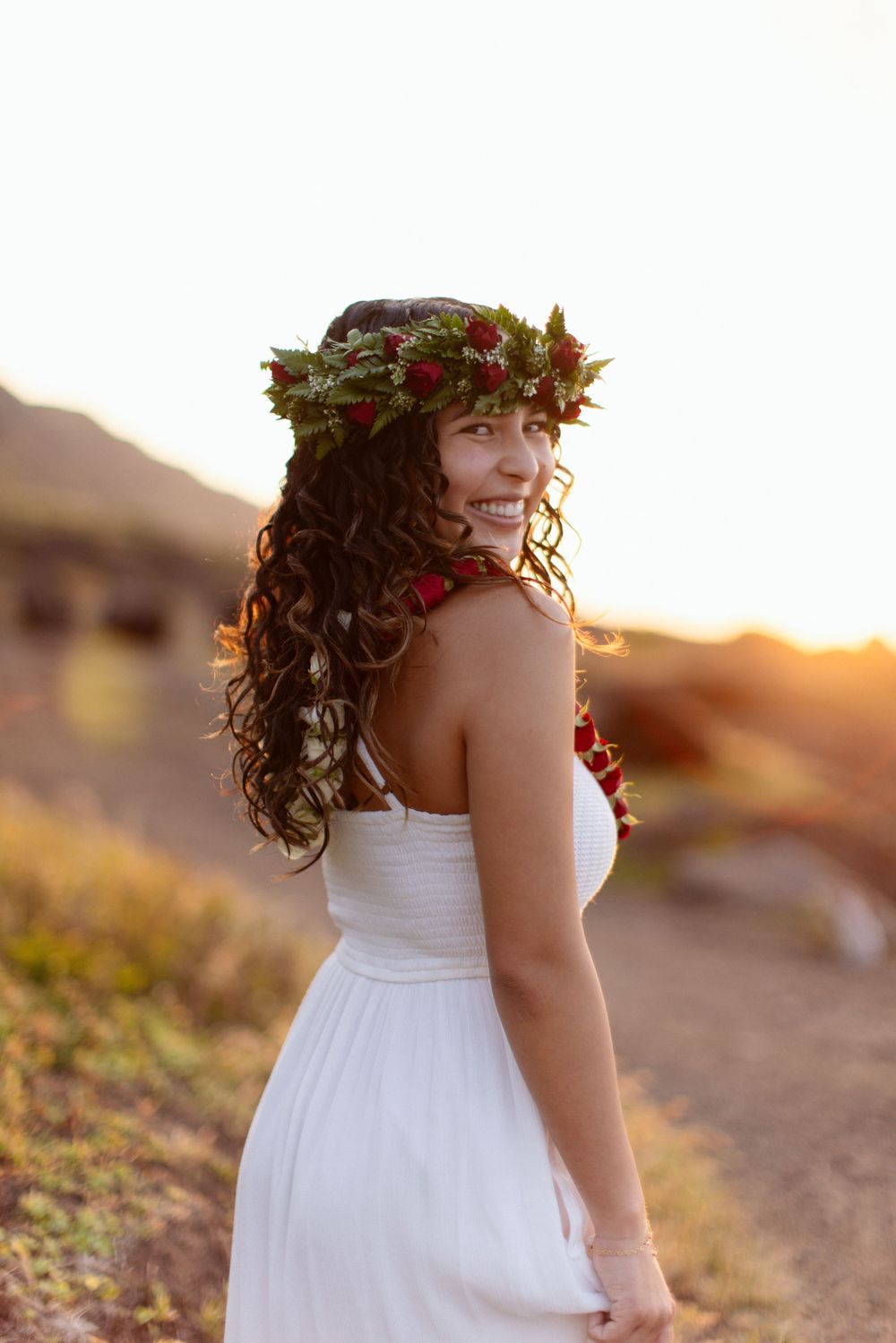 Person in a white dress and floral crown poses at golden hour on a mountain trail.