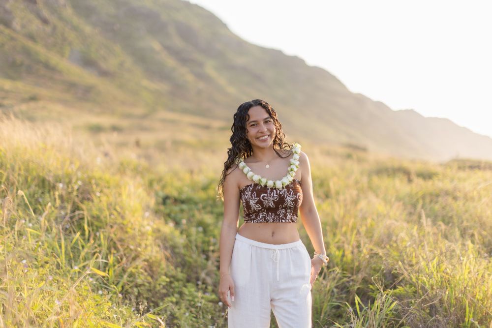 Person in crop top and white pants standing in sun-lit grassy field with mountain landscape in background.