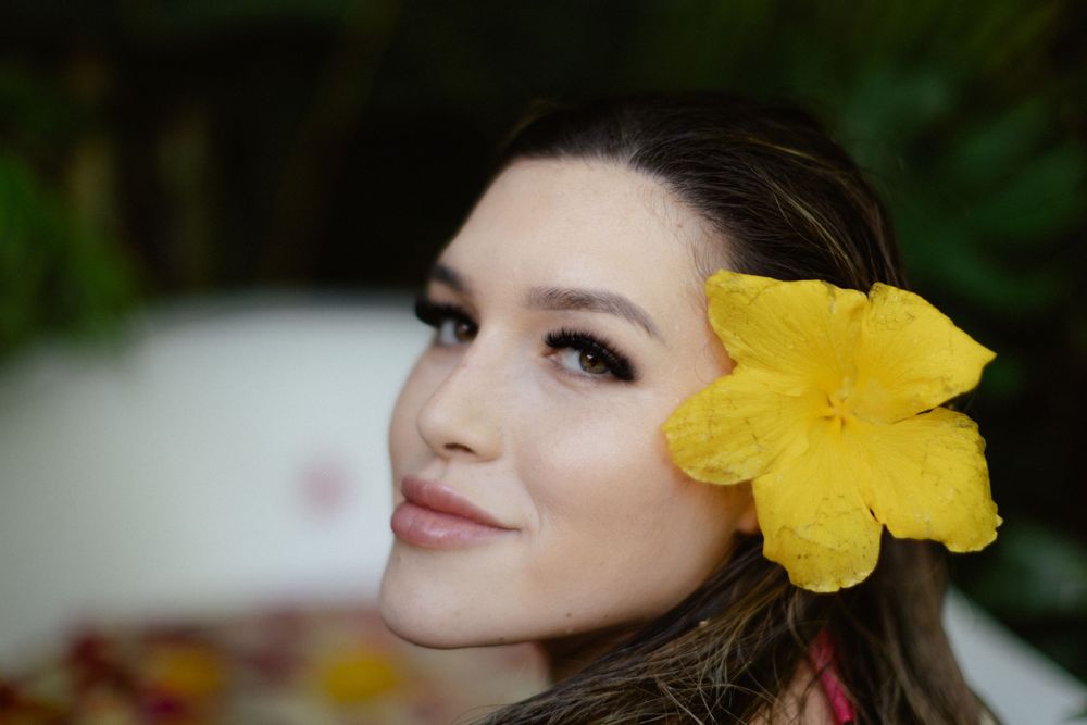 Close up portrait with a bright yellow hibiscus flower worn in long dark hair against a blurred background.