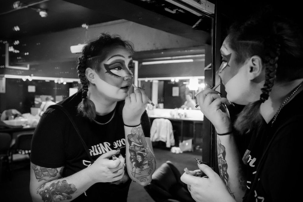 Two people in dark clothing apply makeup backstage in moody black and white lighting.