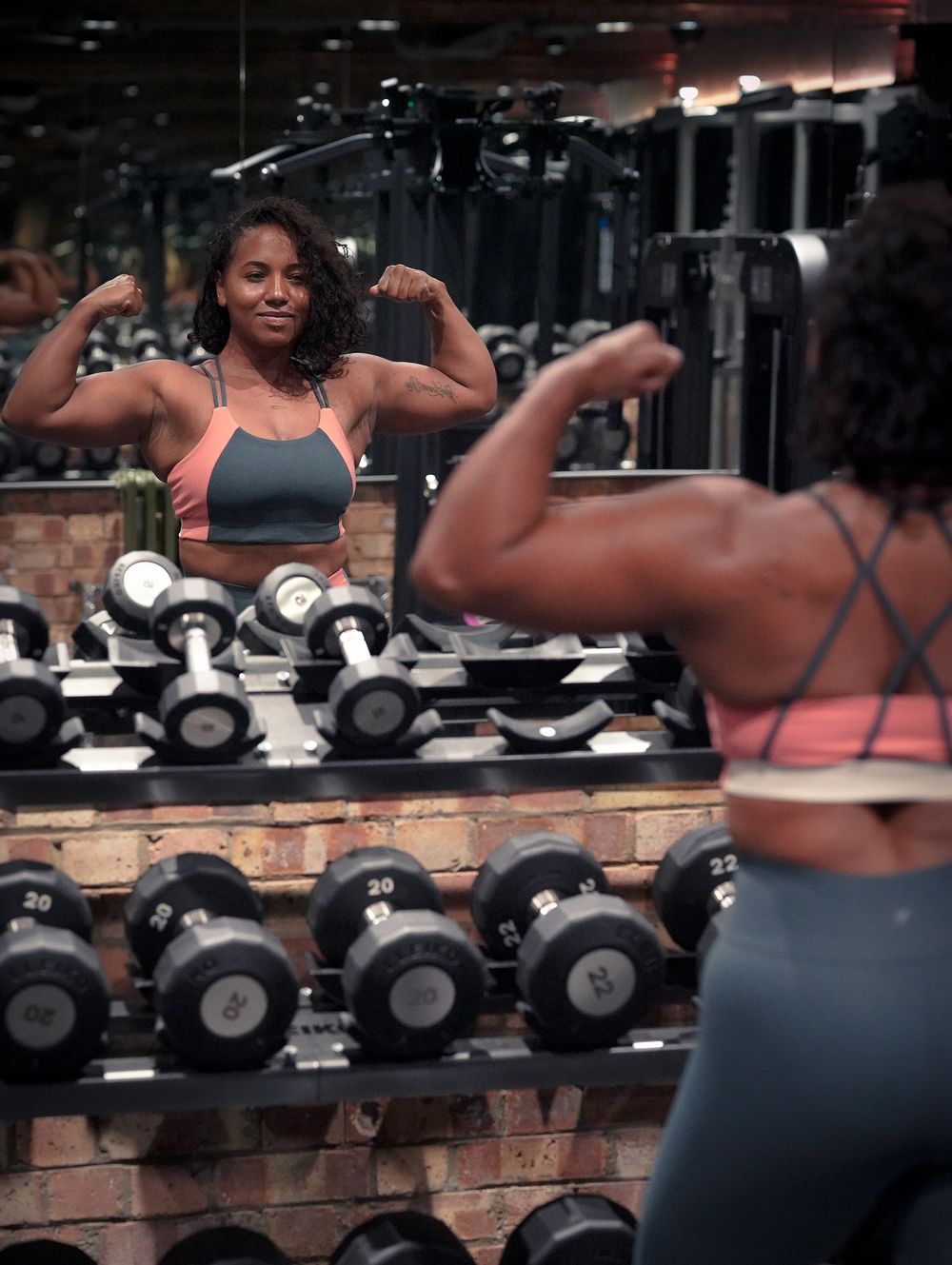 Athletic person flexing muscles while looking in gym mirror with dumbbells in background.