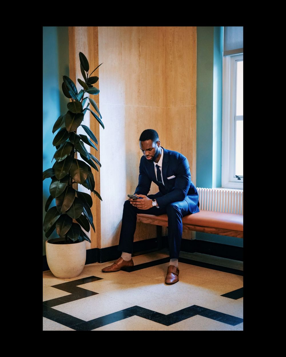 Business professional in suit checking phone while seated next to large indoor plant.