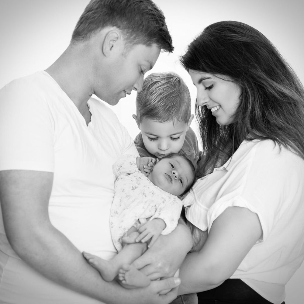 zwart wit foto van newbornshoot, gefotografeerd in maas en waal. gezin vader en moeder aan zijkant, broertje in het midden, kusje aan baby