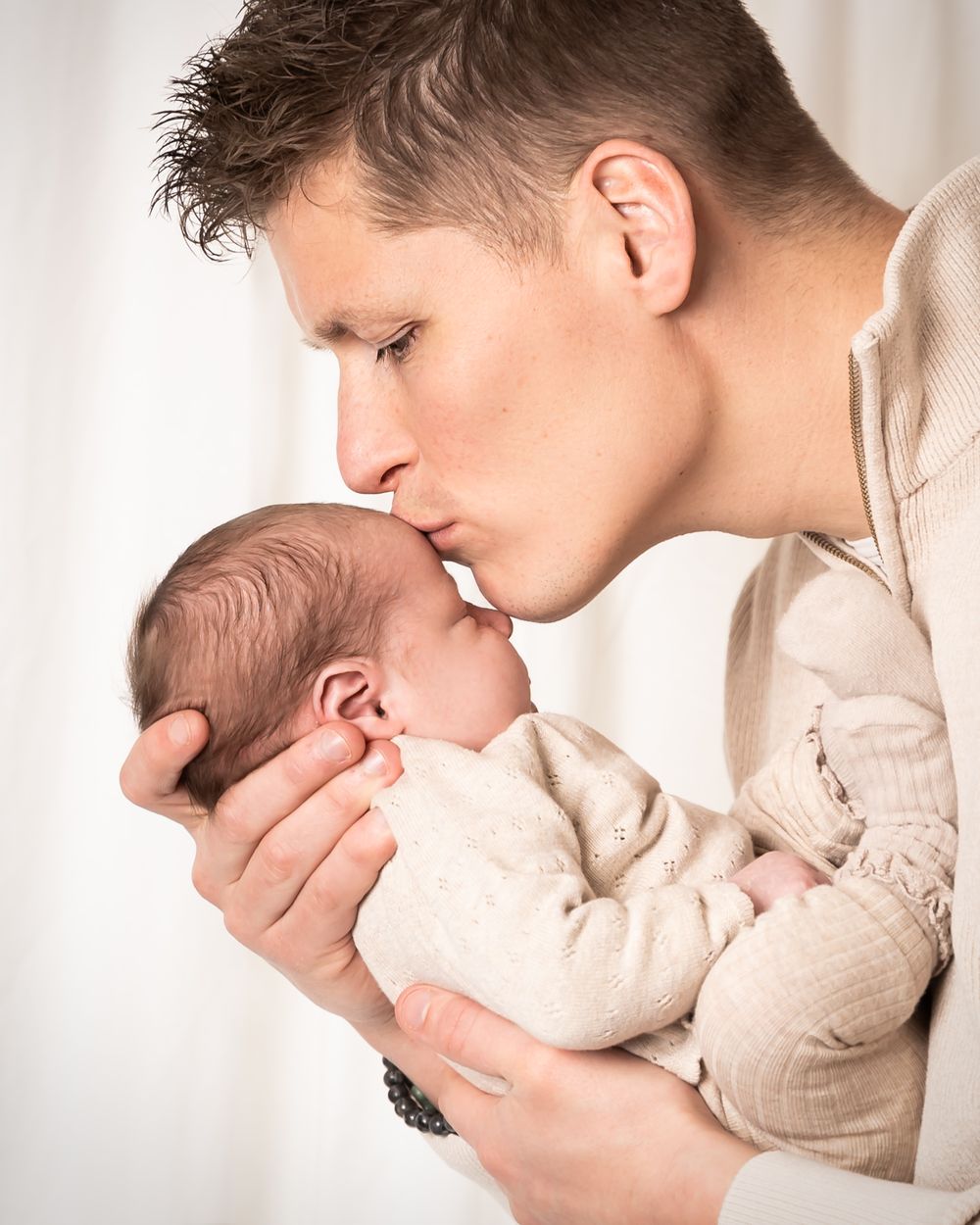 newbornshoot van een pasgeboren baby, vader houdt baby vast en geeft kusje op het voorhoofd, in warme zachte tinten in een studio maas&waal