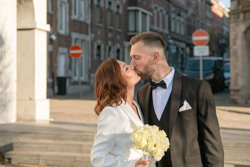 Couple de mariés devant la maison communale en couleur, prêts à se dire oui, échangeant un baiser avec un bouquet de fleurs blanches