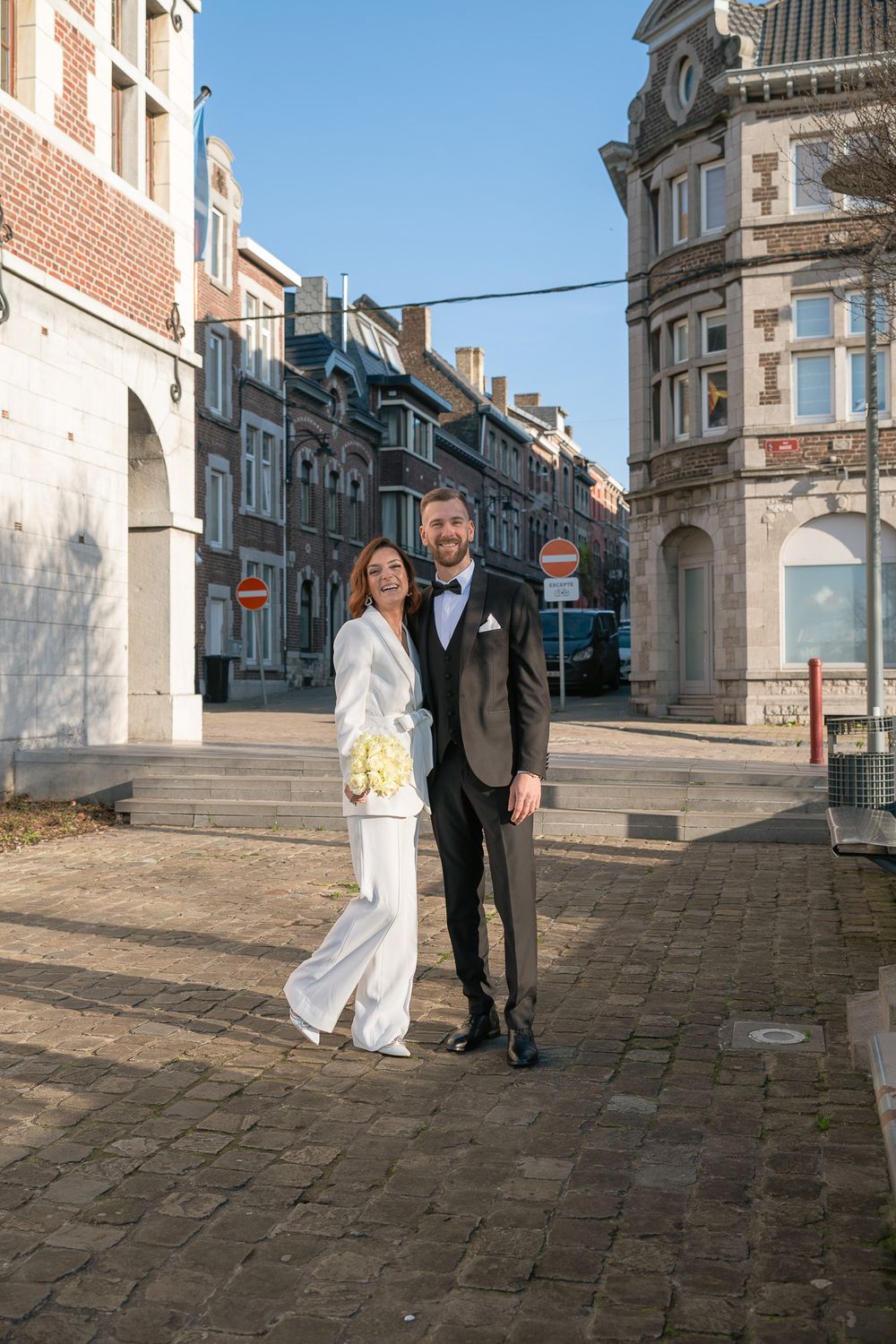Couple de mariés devant la maison communale de Huy sur une rue pavée, bâtiments historiques en arrière-plan, photo en plein soleil