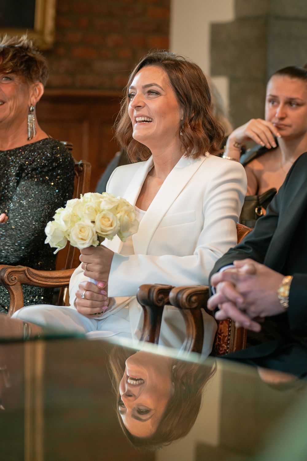 Mariée assise dans la salle communale, souriante, prête à dire oui, tenant un bouquet de roses crème sur une table réfléchissante