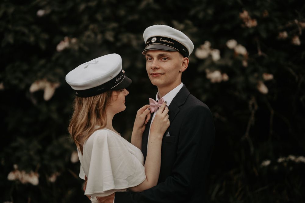 Romantic portrait of couple in naval graduation attire against dark foliage.