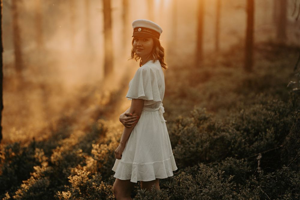 Silhouette of person in white dress and sailor cap standing in sunlit forest.